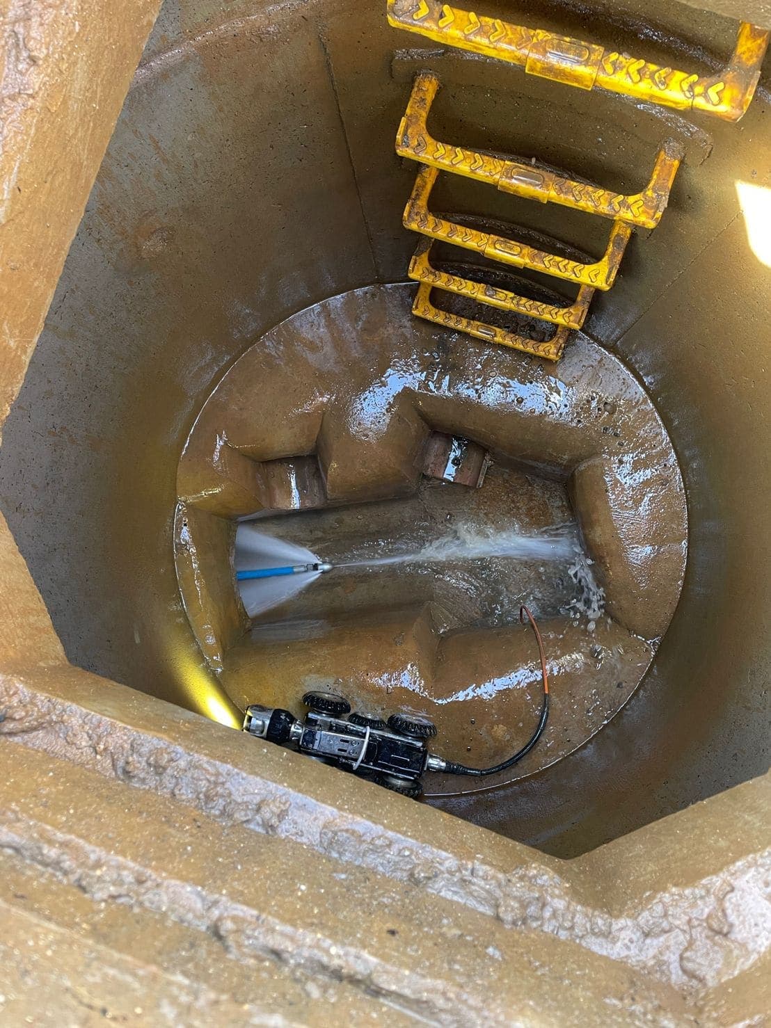 High-pressure jetting nozzle firing across the channel of a manhole chamber, with the CCTV crawler camera at the invert
